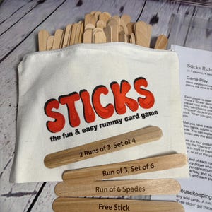 May include: A white canvas bag with a red "STICKS" logo and the text "the fun & easy rummy card game". The bag contains wooden sticks with the text "2 Runs of 3, Set of 4", "Run of 3, Set of 6", "Run of 6 Spades", and "Free Stick".