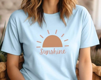 May include: A light blue t-shirt featuring a brown sun graphic with the word "Sunshine" in a cursive font. The shirt is made of a soft material and is being worn by a person sitting in a wicker chair.