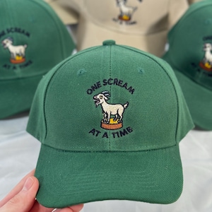 Peut inclure: Casquette de baseball verte avec l'inscription brodée "ONE SCREAM AT A TIME" et un dessin de chèvre. Plusieurs casquettes sont visibles, certaines vertes et d'autres beiges. La casquette est tenue par une main.