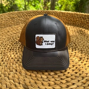 May include: A black and brown trucker hat with a mesh back and a curved brim. The hat features a patch with a cartoon squirrel and the text "What was I doing?"