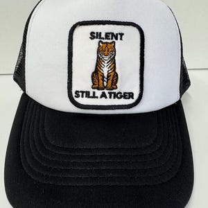 Könnte beinhalten: Weiß-schwarze Trucker-Kappe mit einem schwarzen und weißen gestickten Aufnäher mit der Aufschrift "Silent Still A Tiger" und einem Tiger.