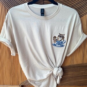May include: A beige t-shirt with a cat wearing glasses sitting in a teacup embroidered on the front.