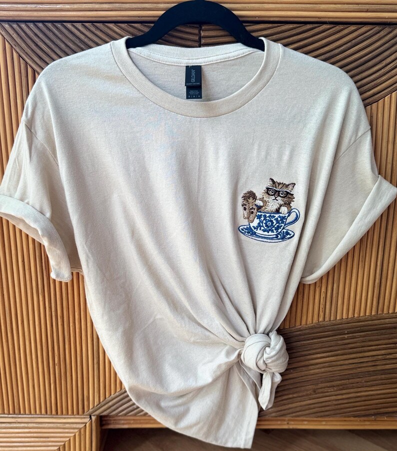 May include: A beige t-shirt with a cat wearing glasses sitting in a teacup embroidered on the front.