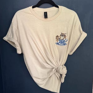 May include: A beige t-shirt with a cat wearing glasses sitting in a teacup embroidered on the front.