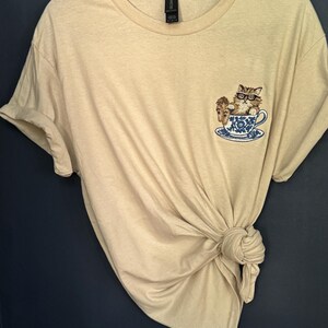 May include: A beige t-shirt with a cat wearing glasses sitting in a teacup embroidered on the front.