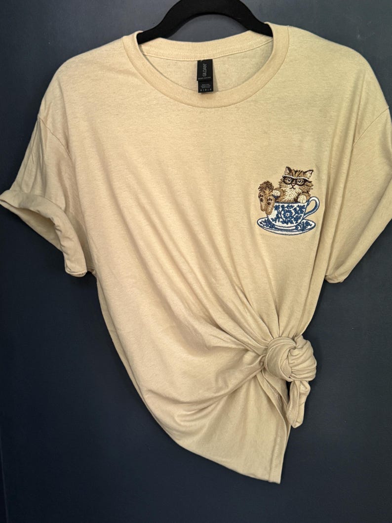 May include: A beige t-shirt with a cat wearing glasses sitting in a teacup embroidered on the front.