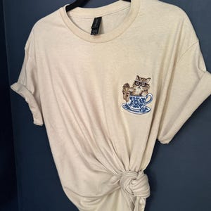 May include: A beige t-shirt with a cat wearing glasses sitting in a teacup embroidered on the front.
