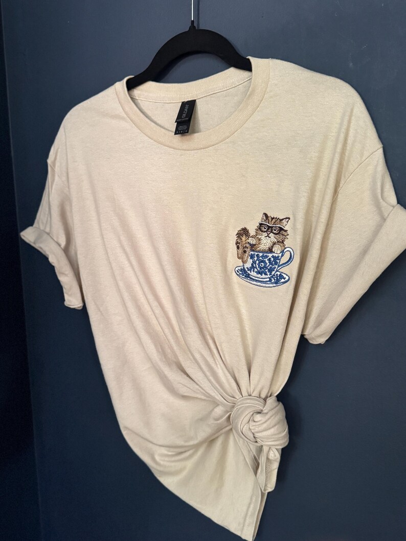 May include: A beige t-shirt with a cat wearing glasses sitting in a teacup embroidered on the front.