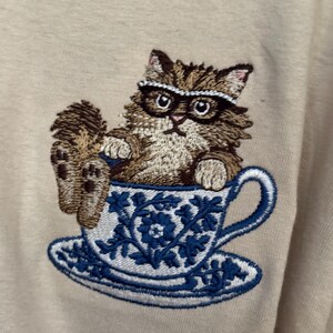 May include: Embroidered design of a brown cat wearing glasses sitting in a blue and white teacup with a saucer.