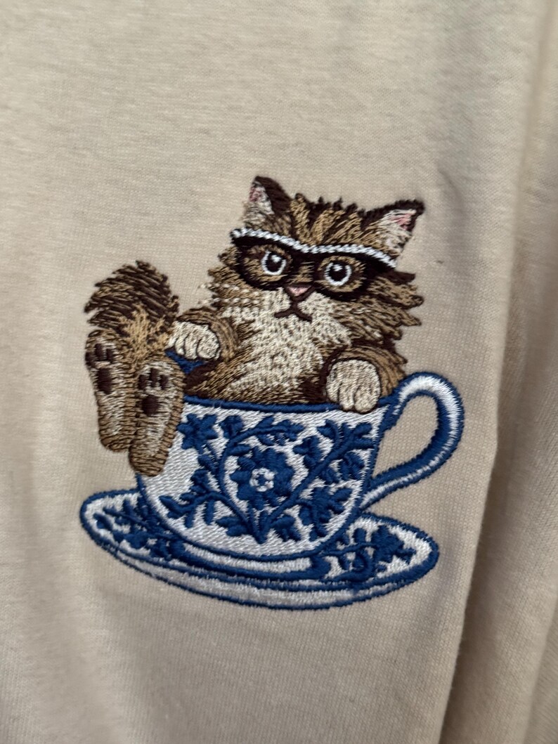 May include: Embroidered design of a brown cat wearing glasses sitting in a blue and white teacup with a saucer.