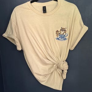 May include: A beige t-shirt with a cat wearing glasses sitting in a teacup embroidered on the front.