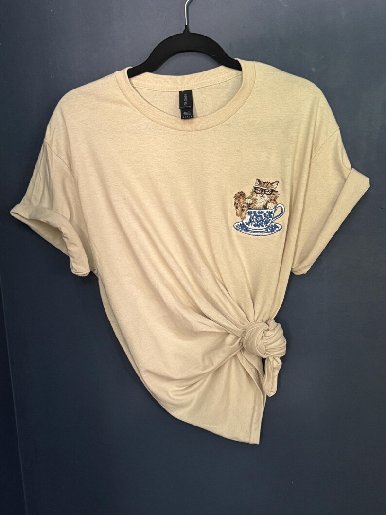 May include: A beige t-shirt with a cat wearing glasses sitting in a teacup embroidered on the front.