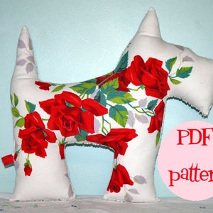 Puede incluir: Una almohada de perro de tela blanca con un patrón de rosas rojas. La almohada tiene forma de Scottish Terrier y tiene una etiqueta de patrón PDF.