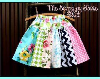 May include: A colorful patchwork skirt with a variety of floral, geometric, and polka dot patterns. The skirt is hanging on a wooden hanger and has a blue, green, pink, white, and black color scheme. The text "The Scrappy Jane Skirt" is written above the skirt.