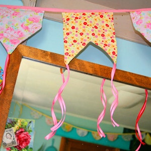 May include: A pink and yellow floral fabric bunting with pink ribbon streamers hanging from a wooden shelf. The bunting is made of fabric with a floral pattern and is hung with pink ribbon streamers. The bunting is hanging in front of a mirror.