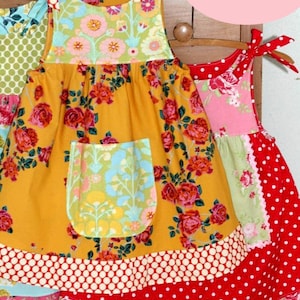 May include: A yellow, pink and red patchwork dress with a floral print and a pocket. The dress has a ruffled hem and is made of cotton fabric. The dress is called "Emie Dress".