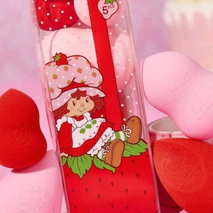 May include: A set of five makeup sponges in a clear box with a red ribbon. The box features a Strawberry Shortcake design. The sponges are pink and red, with a strawberry theme. The packaging includes the number "5" and the word "PCS".