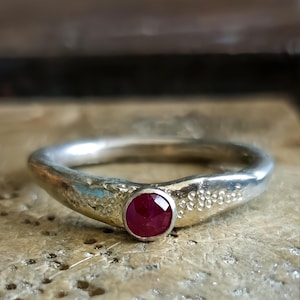 May include: A silver ring featuring a round, faceted, deep red ruby gemstone set in a bezel. The band has a textured, uneven surface with small, raised circular details on either side of the central stone, creating a distinct, organic appearance.