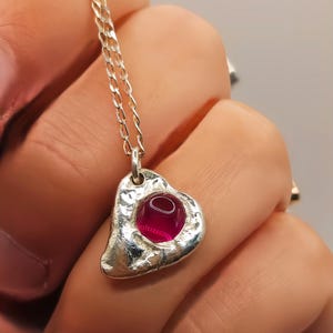May include: A silver-toned necklace featuring a heart-shaped pendant with a textured, hammered surface. A smooth, round, deep pink gemstone is set in the centre of the pendant. The pendant hangs from a delicate, thin silver-toned chain link necklace.