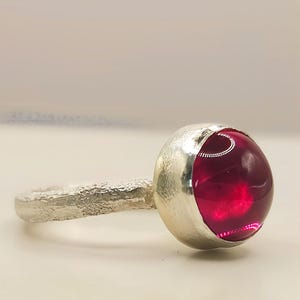 May include: A silver ring featuring a round, deep pink cabochon gemstone set in a smooth bezel. The ring band has a textured, matte finish that contrasts with the polished metal of the setting and the glossy, translucent surface of the stone.