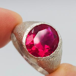 May include: A ring featuring a large, oval-cut, vibrant red gemstone set in a textured, matte silver-toned metal band. The stone has multiple facets that reflect light, while the band displays a granular, sandblasted finish around the setting.