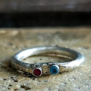 May include: A silver ring with a textured, hammered finish featuring two small, round, bezel-set gemstones. One stone is a deep red colour, and the other is a bright blue colour. The band has an irregular, organic shape with a metallic, reflective surface.