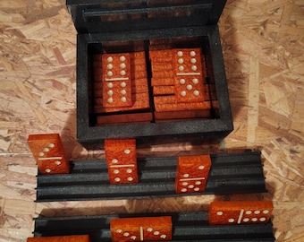 Puede incluir: Un juego de dominó de color naranja-marrón con puntos blancos, guardado en una caja rectangular negra. El juego incluye dos soportes de plástico negro que sostienen varias piezas en posición vertical, mostrando diversas combinaciones de puntos.