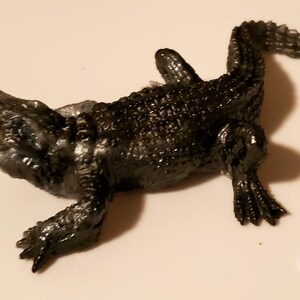 May include: A black plastic toy alligator with a textured surface. The alligator is lying on its back with its legs and tail outstretched.