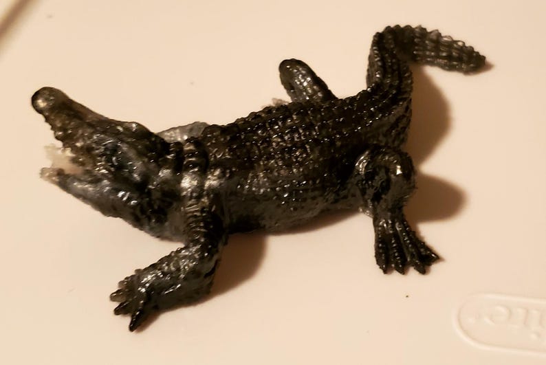May include: A black plastic toy alligator with a textured surface. The alligator is lying on its back with its legs and tail outstretched.