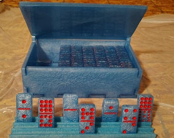 Puede incluir: Un juego de dominó azul con puntos rojos, exhibido en un soporte azul escalonado. El juego incluye una caja azul a juego con tapa con bisagras, llena de fichas de dominó adicionales. Los dominós están dispuestos en fila.