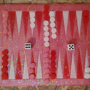 Puede incluir: Un tablero de backgammon rosa con puntas triangulares rojas y blancas. Las fichas rojas y blancas están dispuestas en el tablero, junto con dos dados que muestran los números seis y cinco. El tablero tiene un patrón de corazón.