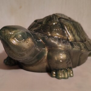 May include: A green and gold turtle ornament. The figurine has a detailed shell and is in a resting pose. The glossy material gives it a polished look. The ornament is suitable for display.