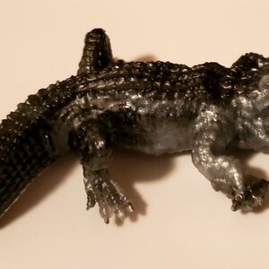 May include: A black and silver toy alligator with an open mouth and textured scales. The alligator is posed with its head raised and its tail curled.