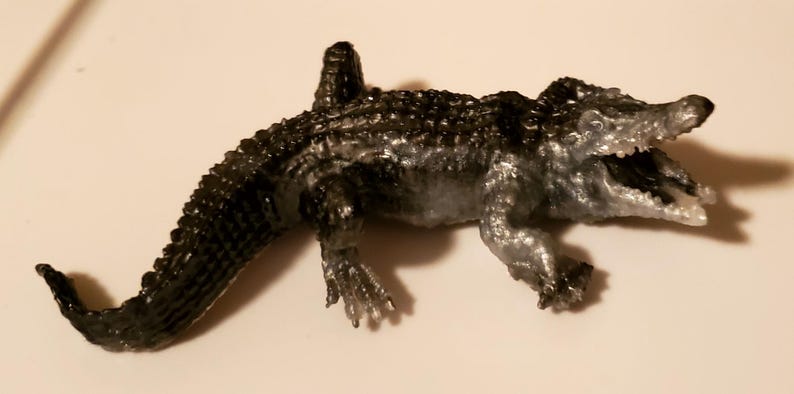May include: A black and silver toy alligator with an open mouth and textured scales. The alligator is posed with its head raised and its tail curled.