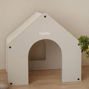 以下が含まれることがあります： 切妻屋根のデザインとアーチ型の入り口を持つ白いペットハウス。前面パネルには小文字の白い文字でharleyという名前が印刷されています。構造は角と頂点にある目に見える暗い円形の留め具で組み立てられています。