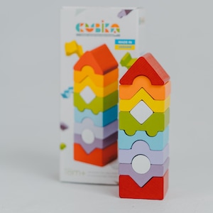 Puede incluir: Un colorido juguete de apilamiento de madera con forma de casa. El juguete está hecho de bloques de varios colores del arcoíris con recortes blancos en forma de círculo y diamante. La caja detrás del juguete tiene la palabra "cubika" impresa.