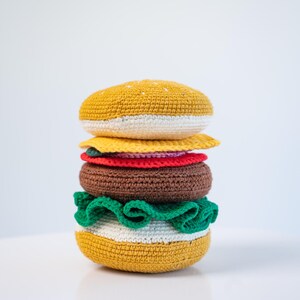 May include: A crocheted burger with yellow, white, brown, red, and green components. The top and bottom buns are yellow with white trim. The burger includes a brown patty, yellow cheese, red tomato, and green lettuce.
