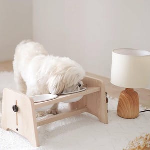 May include: A light-colored wooden elevated pet feeder with two white ceramic bowls. A small white dog is eating from one of the bowls. A wooden lamp with a cream-colored shade sits to the right.