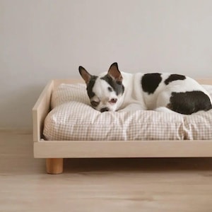 Puede incluir: Un perro pequeño de color blanco y negro descansa sobre una cama rectangular de madera clara para mascotas. La cama cuenta con un marco bajo con patas cortas y está cubierta por un cojín suave con un patrón de tela de cuadros vichy beige y blanco.