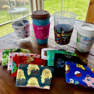May include: A collection of colorful fabric coffee cup sleeves with various designs, including a pink sleeve with the word "Barbie" on it, a blue sleeve with a Pokemon character, and a green sleeve with a "What Would Poof Do?" design.