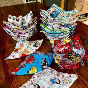 May include: A collection of colorful fabric bowl cozies with various patterns, including Disney characters and movie themes. The cozies are stacked and displayed on a wooden surface, showcasing a variety of designs and colors.