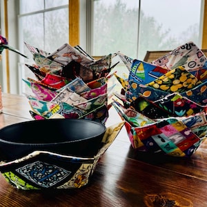 May include: A set of colorful fabric bowl cozies, made from different patterned fabrics, with a black bowl inside one of the cozies. The cozies are folded into a bowl shape and are stacked on a wooden table.