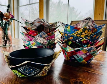 May include: A set of colorful fabric bowl cozies, made from different patterned fabrics, with a black bowl inside one of the cozies. The cozies are folded into a bowl shape and are stacked on a wooden table.