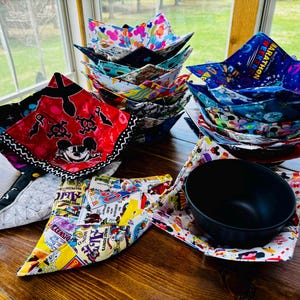 May include: A collection of colorful fabric bowl cozies with various Disney character designs, including Mickey Mouse, pirates, and a "RunDisney" marathon theme. A black bowl is sitting inside one of the cozies.