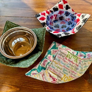 May include: Three bowl cozies with different patterns. One is green with a small pattern, another has a playing card design, and the third features the word "IOWA" and other text. The bowls are on a wooden surface.