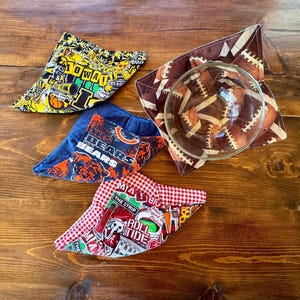 May include: Four fabric bowl cozies featuring various sports team prints including Iowa, Chicago Bears, and Alabama themes. One cozy contains a clear glass bowl with a brown football pattern. The fabric designs include team logos, text, and colors.