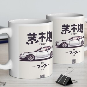 Może przedstawiać: Biały kubek ceramiczny z ilustracją białego samochodu i japońskim tekstem, który brzmi "Kyoto" i "2,5". Samochód to białe coupe z czarnymi akcentami. Kubek ma czarny uchwyt.