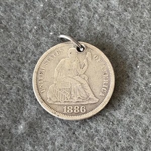 May include: A silver coin pendant from 1886 featuring a seated Liberty design. The coin is attached to a silver loop. The words "UNITED STATES OF AMERICA" are visible around the edge of the coin.