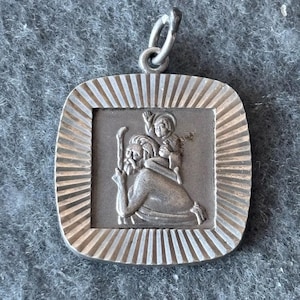 Könnte beinhalten: Silberner Anhänger mit einem detaillierten Relief von Sankt Christophorus, der ein Kind auf seinen Schultern trägt. Der Anhänger ist quadratisch mit einem dekorativen Rand und einer Schlaufe zum Aufhängen.