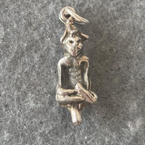 Può includere: Un ciondolo d'argento a forma di figura seduta con un cappello a punta. La figura ha un viso e delle mani dettagliate.